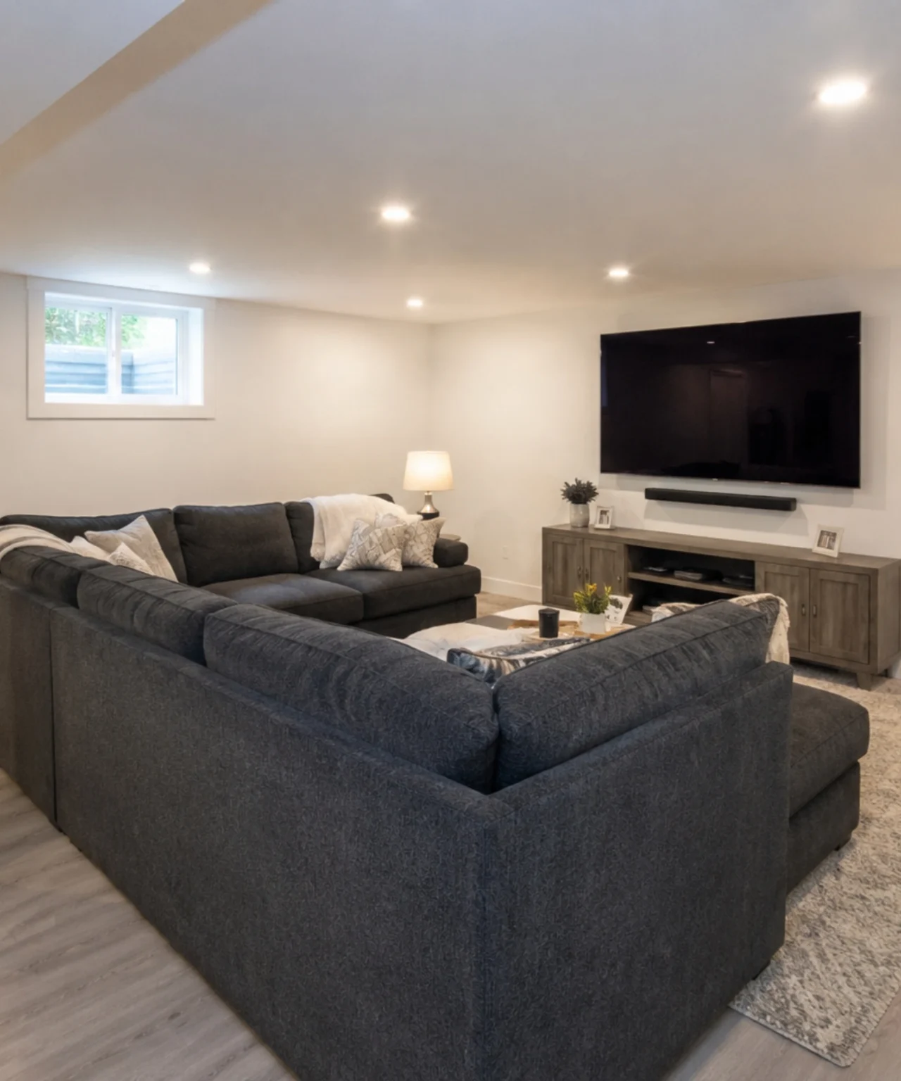 Finished basement media room with charcoal sectional sofa, wall-mounted TV, recessed lighting, and small basement window.